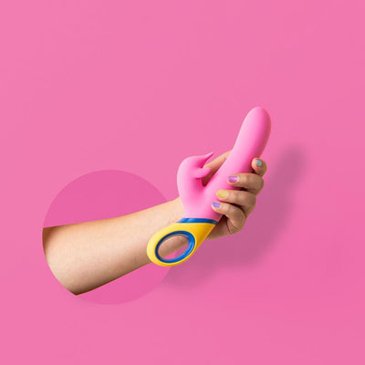 Hand holding a pink silicone dolphin vibrator with yellow and blue handle against a pink background.
