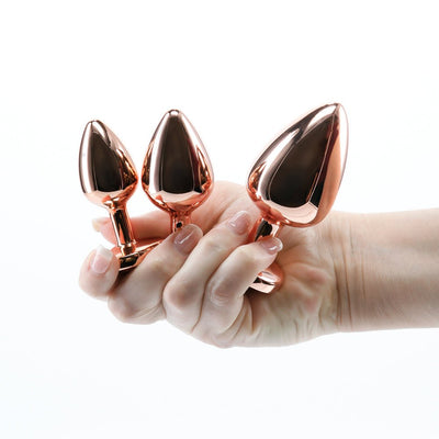 Hand holding three graduated rose gold metal plugs from the Rear Assets 3-piece Trainer Kit against a white background.