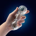 Hand holding a clear textured pleasure toy with ribbed and nodular chambers against a dark blue background.