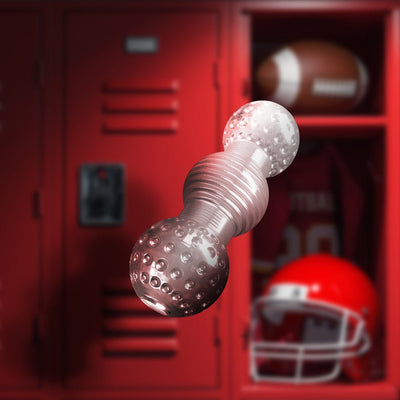 Clear textured dual-ended pleasure toy with ribbed center, shown against a red locker room background with football gear.
