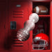 Clear textured dual-ended pleasure toy with ribbed center, shown against a red locker room background with football gear.