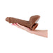 Hand holding a large dual-density realistic silicone dildo with a suction base in brown color.