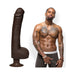 Lifelike dark brown 12-inch dildo with suction base next to shirtless man with tattoos and jewelry against white background.