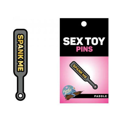 Gray enamel paddle pin with gold "SPANK ME" text, displayed on a pink and black Sex Toy Pins backing card.