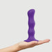 Hand holding a purple silicone dildo with ergonomic curves and a suction cup base against a plain background.
