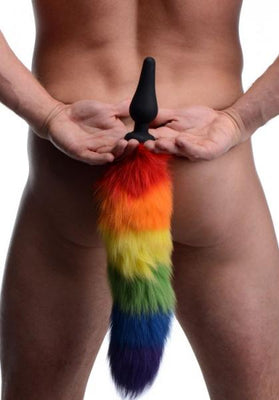 Rear view of a person holding a black silicone butt plug with a long, fluffy rainbow-colored tail attached.