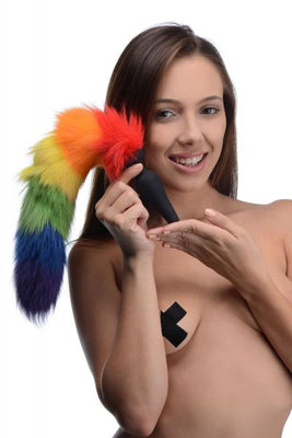 Woman holding a rainbow-colored faux fur tail silicone butt plug, smiling against a white background.