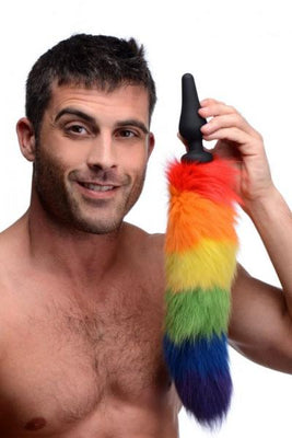 Smiling shirtless man holding a black silicone butt plug with a long, fluffy rainbow-colored faux fur tail.