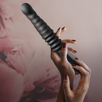 Hands holding a black ribbed thrusting dildo with a curved tip against a soft pink floral background.