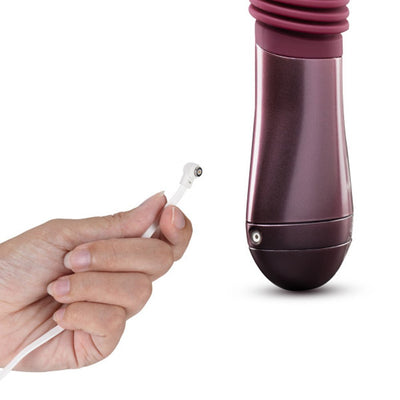 Hand holding magnetic USB charging cable near the base of the Temptasia Trixie thrusting dildo in wine color.