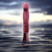 Temptasia Trixie thrusting dildo in wine color standing upright in calm water with a soft cloudy sky background.
