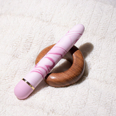 Pink and white marbled vibrator resting on a wooden ring on a soft textured fabric background.