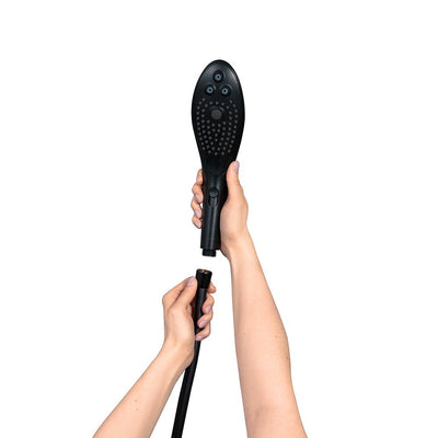 Hands holding and connecting a sleek black Womanizer Wave Shower Head Masturbator with hose attachment on a white background.