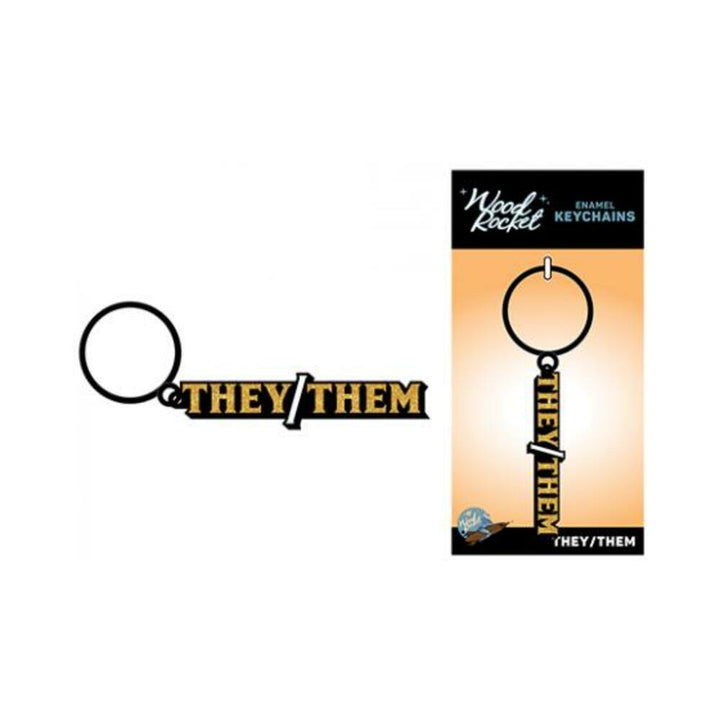 Black and gold enamel keychain with "They/Them" text by Wood Rocket, shown both loose and on branded packaging.